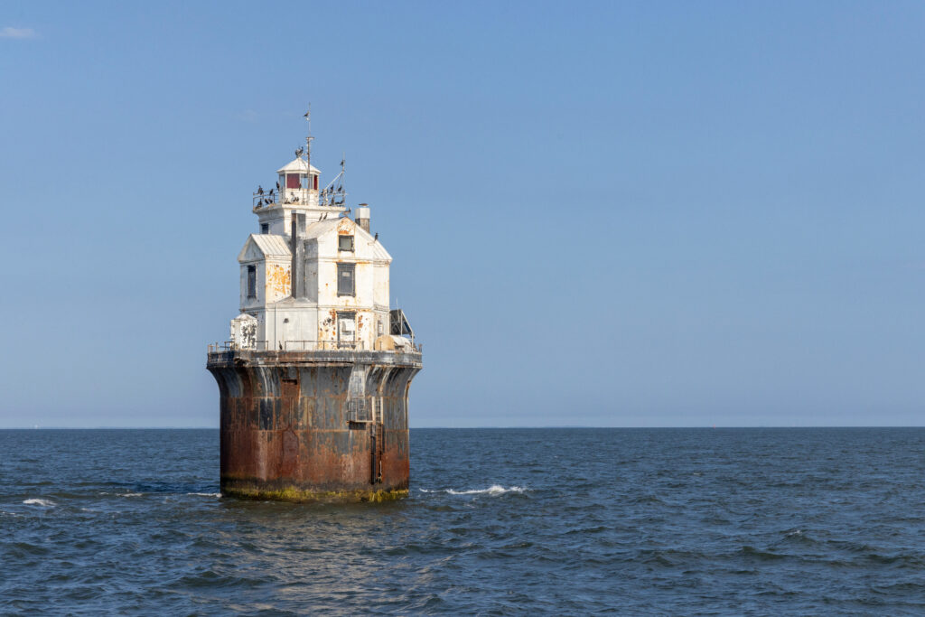 Weathered white lighthouse structure with a rust-stained caisson base standing alone in open water under a clear blue sky.