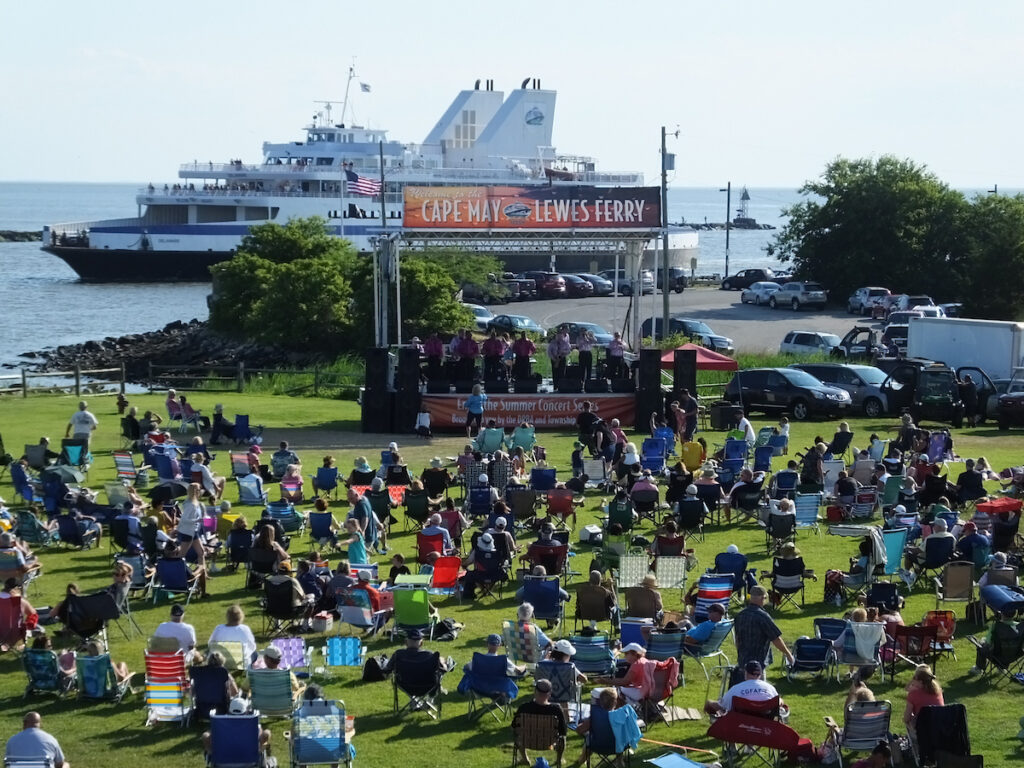 Concert-goers sit on their lawn chairs on the "green" next to the Cape May Ferry terminal in front of the stage, with the Ferry sailing in the background