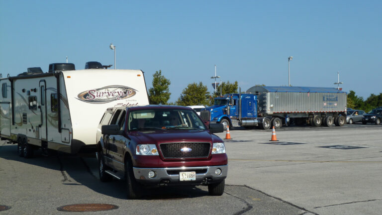 Semi-truck tractor trailer in the background, with cars waiting to load the Cape May - Lewes Ferry with a truck pulling a camper in the foreground driving aboard