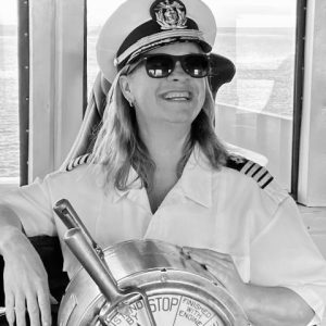 Black and white photo of a female Ferry Captain with her uniform, hat, and sunglasses on as she steers the Ferry